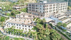 A large, multi-story building with numerous balconies and terraces, surrounded by lush greenery and palm trees. - Hotel Antares (Letojanni)