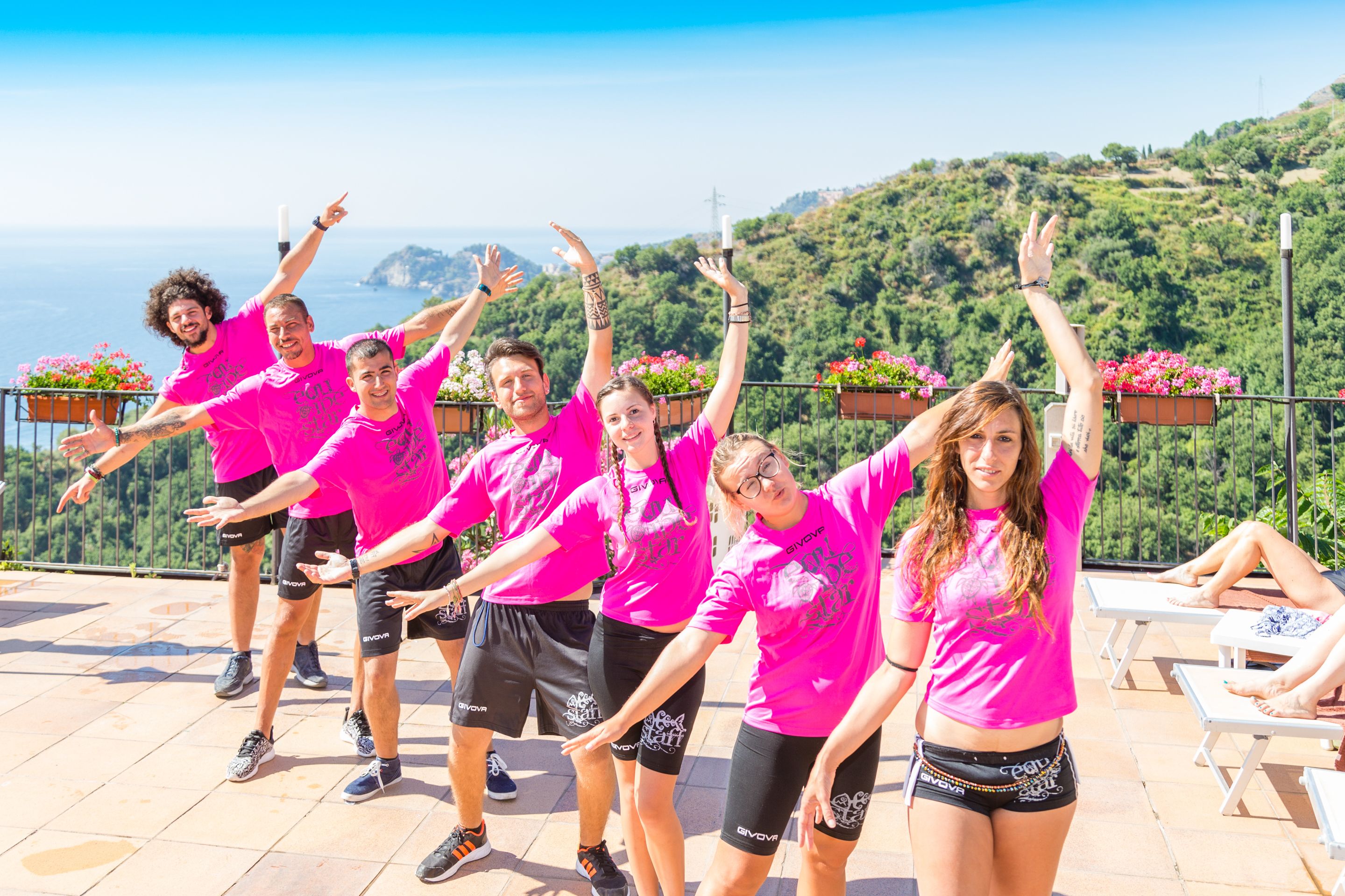 A group of people in pink shirts performing an exercise routine on a terrace with a scenic view of hills and the ocean.