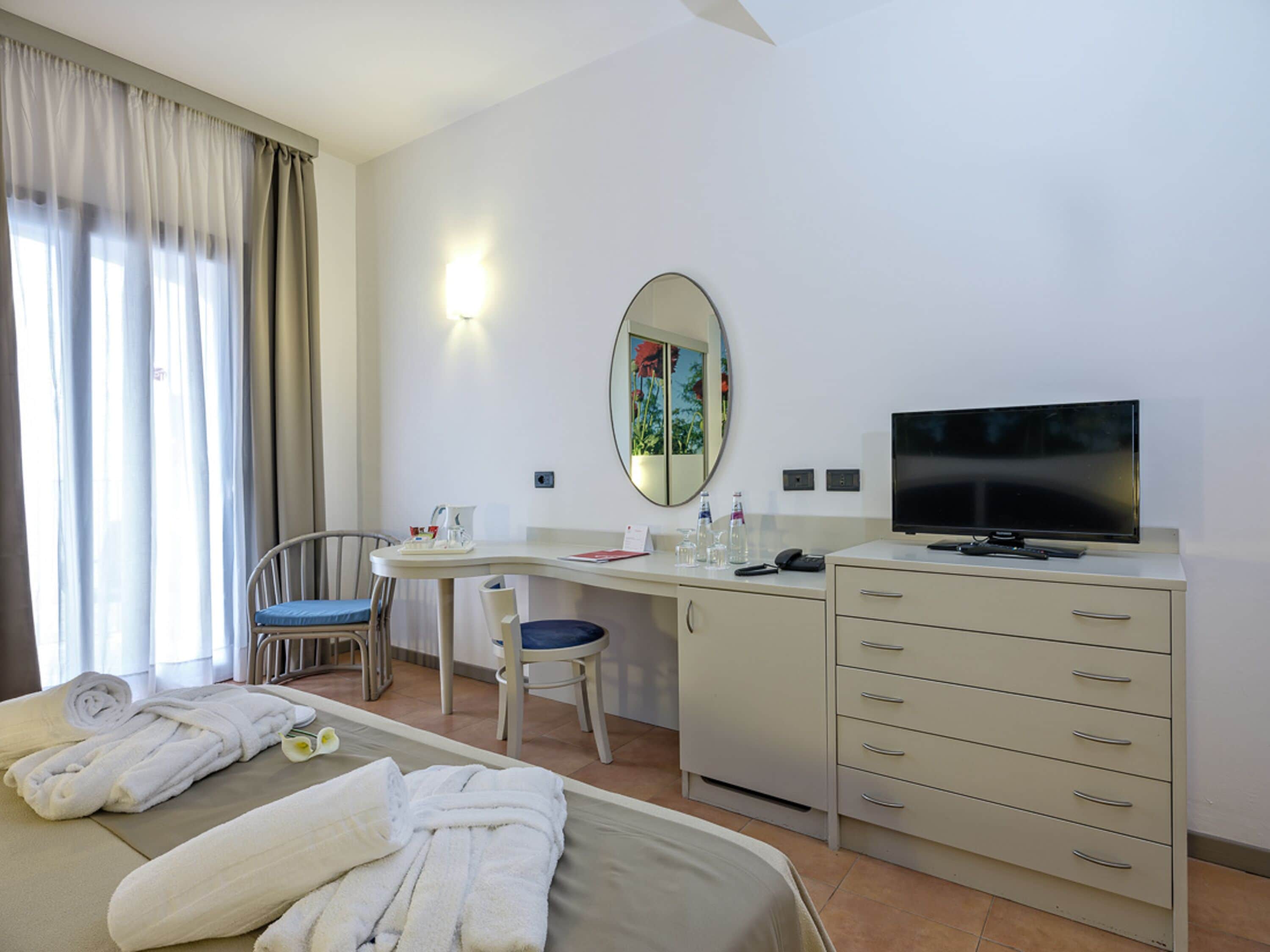 A hotel room with a bed, a desk with a chair, a television, a mirror, and a wardrobe.