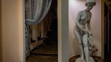 Um corredor com uma estátua de mulher segurando um tecido, uma escadaria ao fundo e uma cortina estampada.