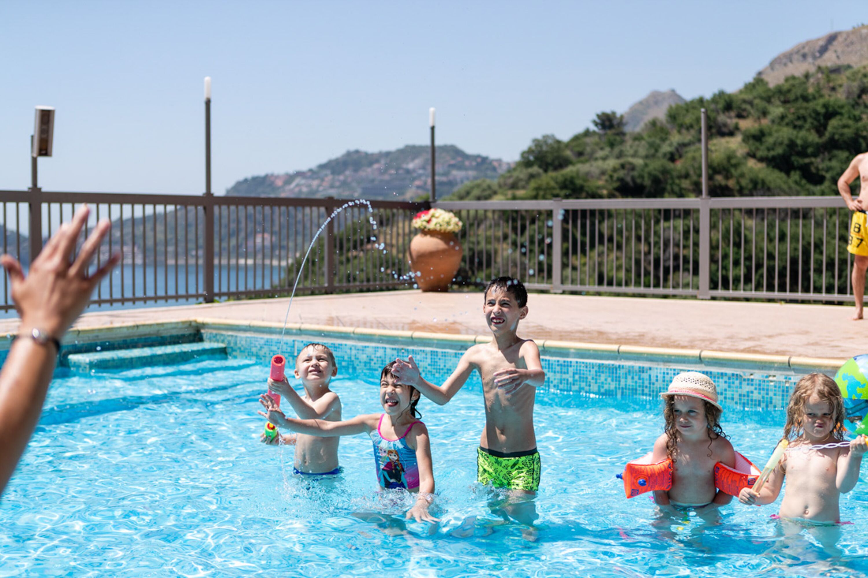 Una piscina con bambini che giocano, un grande vaso da fiori e una vista sulle colline sullo sfondo.