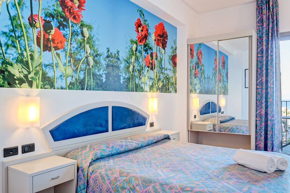 A hotel room with a large bed, a bedside table, a nightstand, a mirror, and a window with curtains.