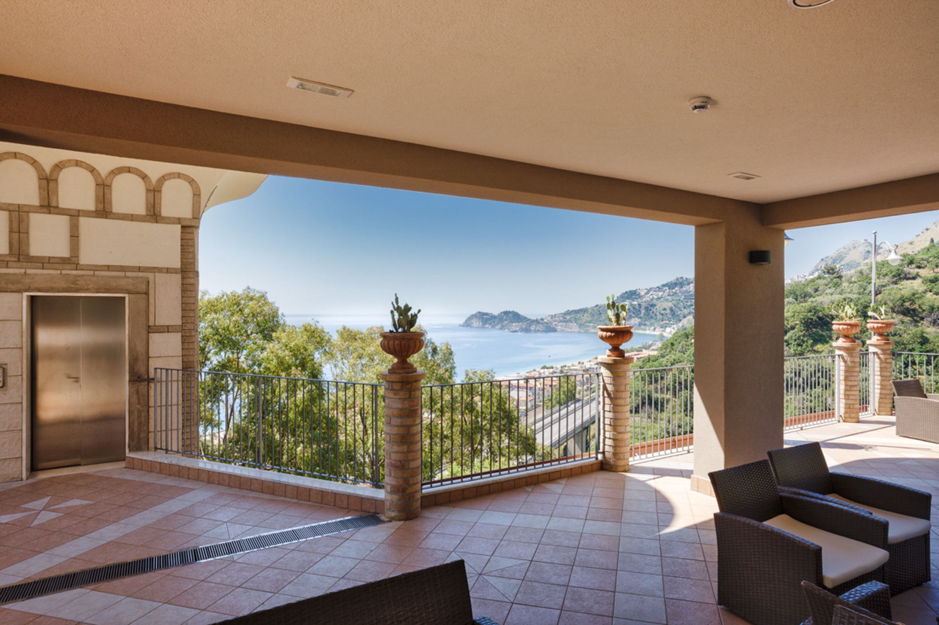 A spacious balcony with a view of the sea and hills, furnished with wicker chairs and a potted plant.