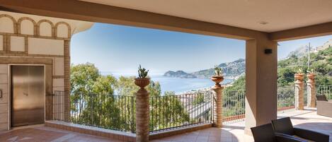 A spacious balcony with a view of the sea and hills, furnished with wicker chairs and a potted plant.