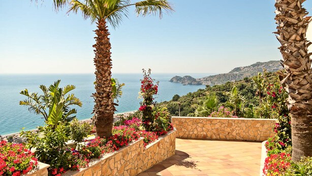 A terrace with a stone wall, palm trees, and colorful flowers. - Hotel Antares (Letojanni)