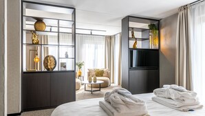Una habitación de hotel moderna con una cama, un televisor, una zona de estar con un sofá y una pequeña mesa, y una estantería decorativa con objetos.