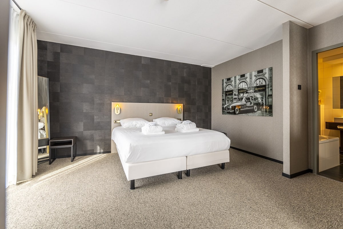 A modern hotel room with a large bed, a small bedside table, a wall-mounted light, and a poster on the wall.