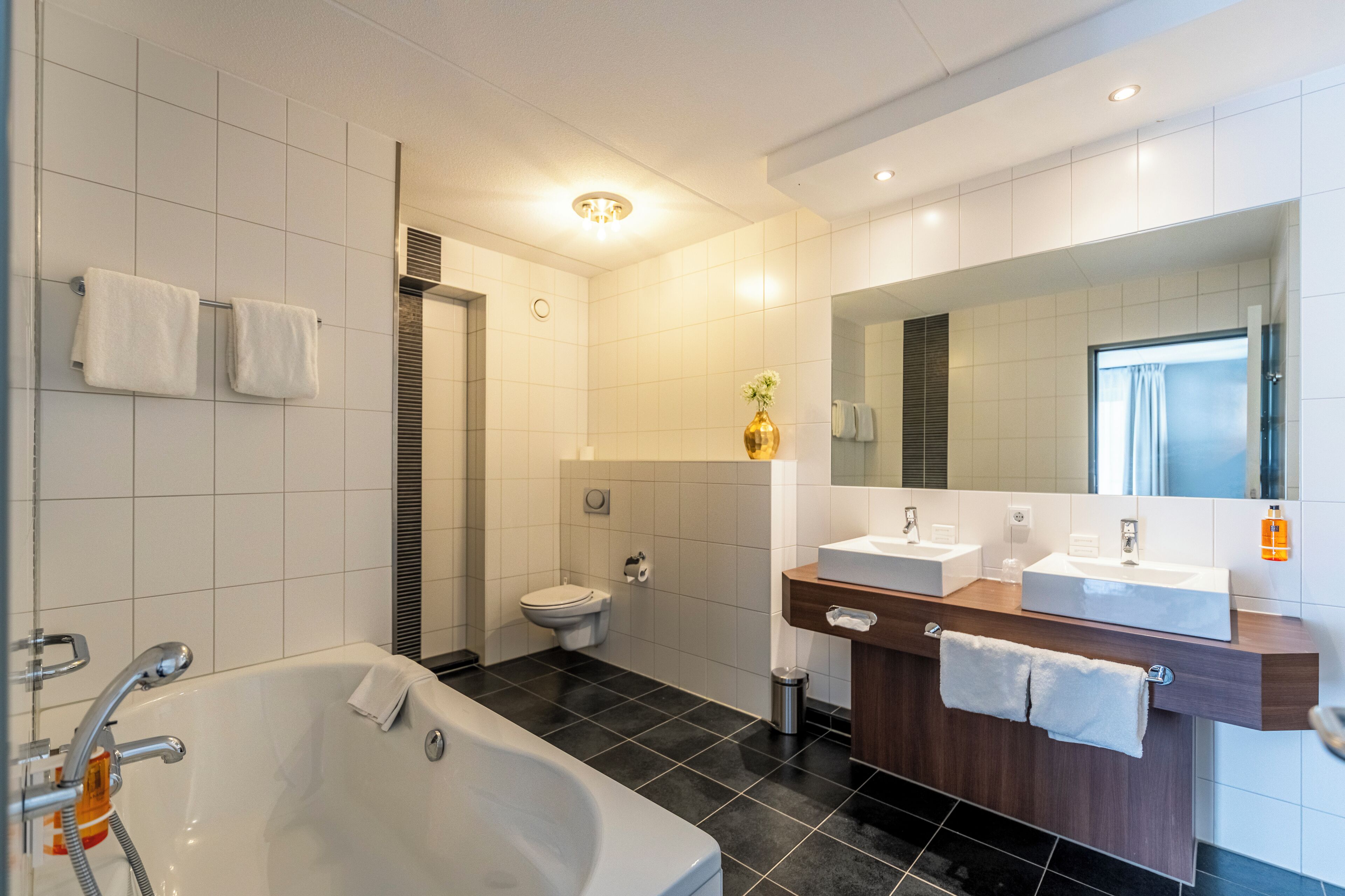 A modern bathroom with a bathtub, two sinks, a toilet, and a large mirror.