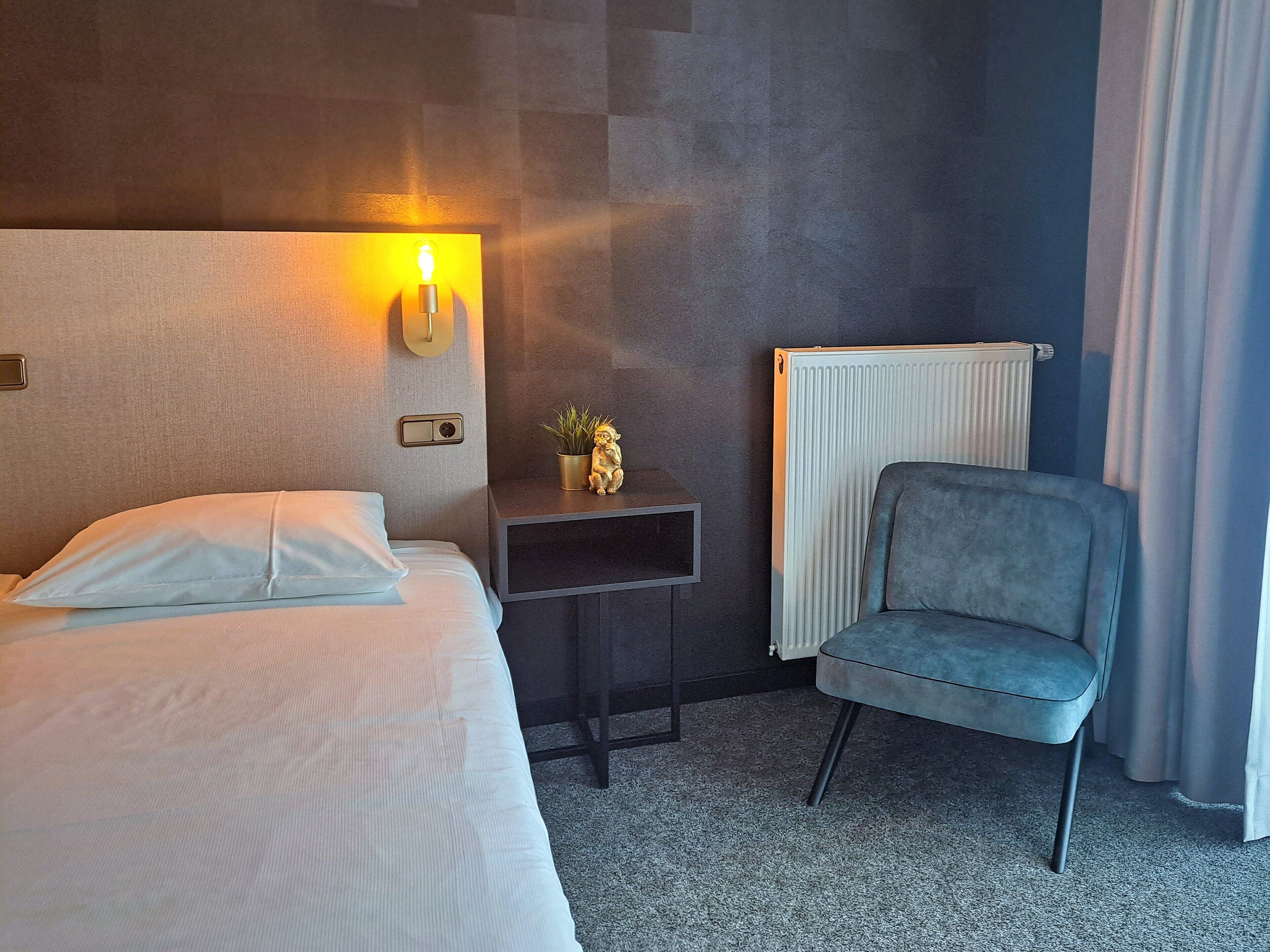 A hotel room with a bed, a small table with a plant, a chair, and a radiator.