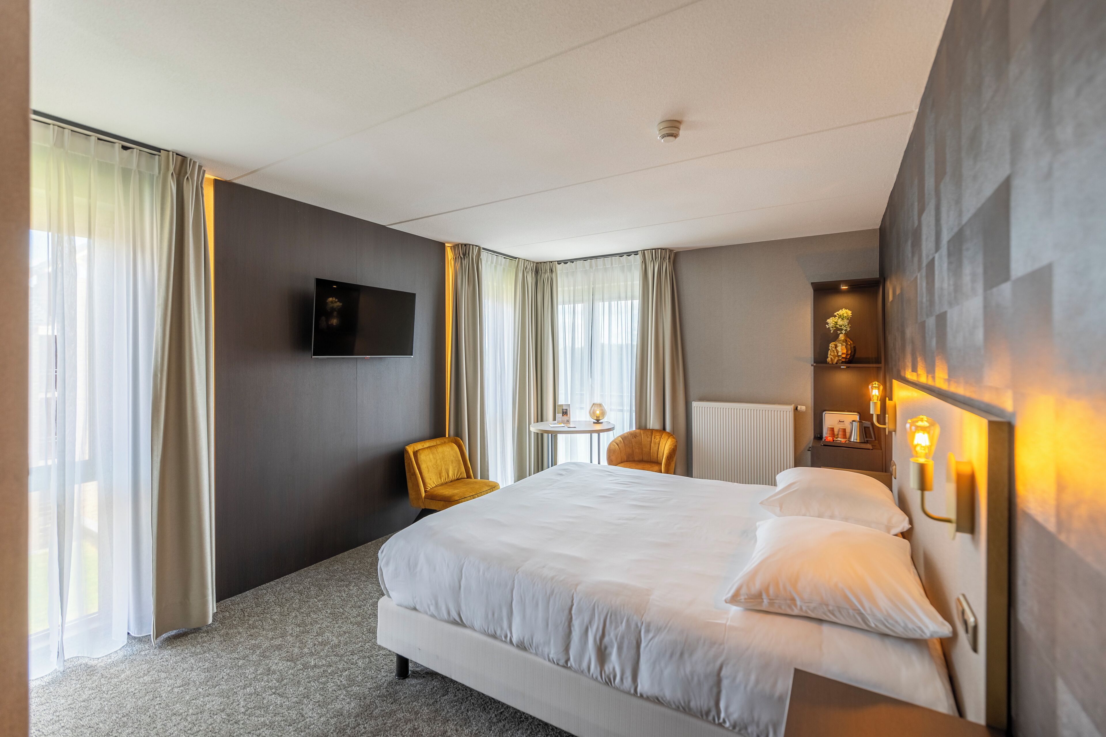 A hotel room with a large bed, two chairs, a small table, and a TV on the wall.