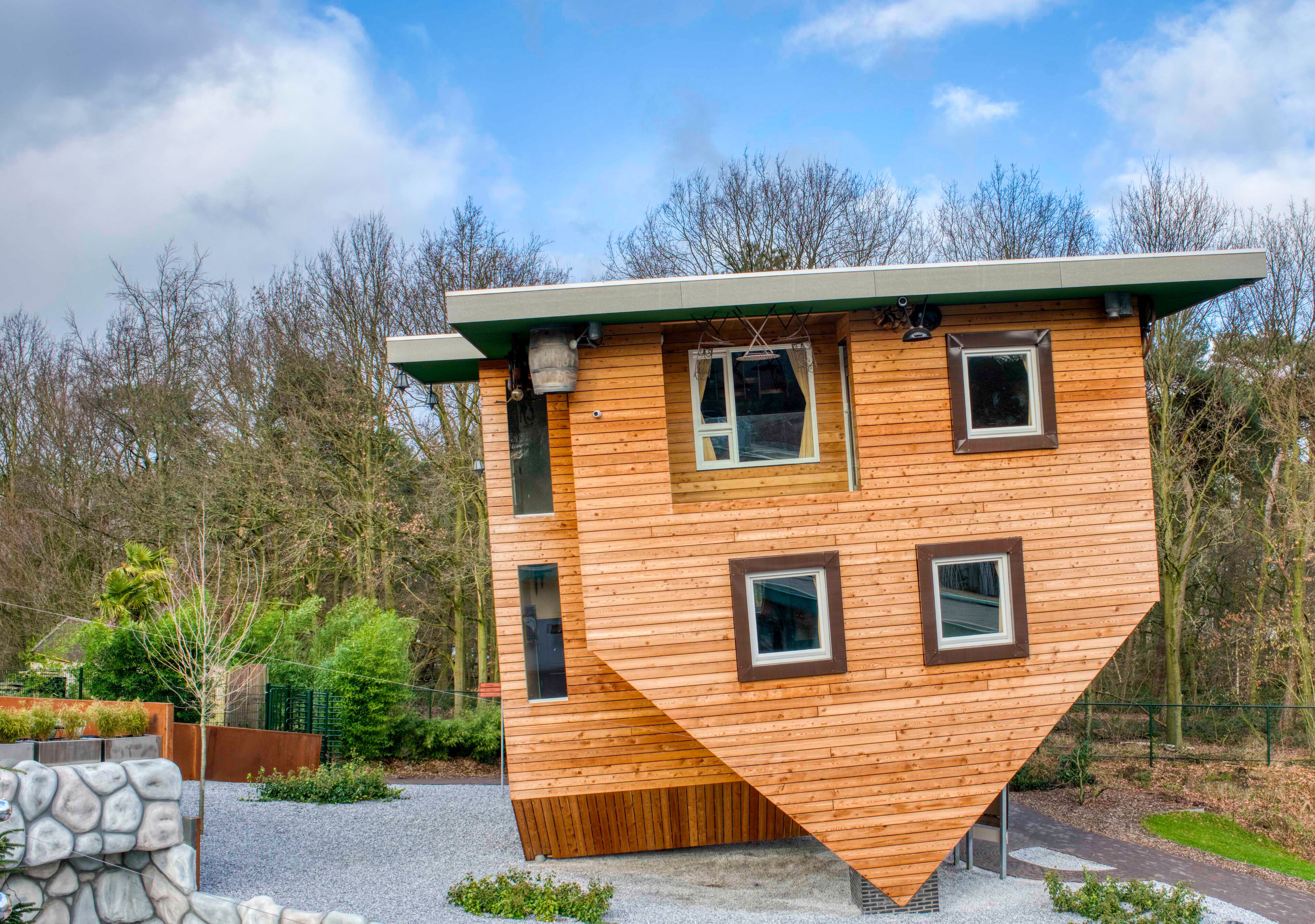 A unique, triangular wooden house with multiple windows and a green roof, surrounded by trees and a gravel path.