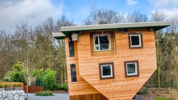 A unique, triangular wooden house with multiple windows and a green roof, surrounded by trees and a gravel path.