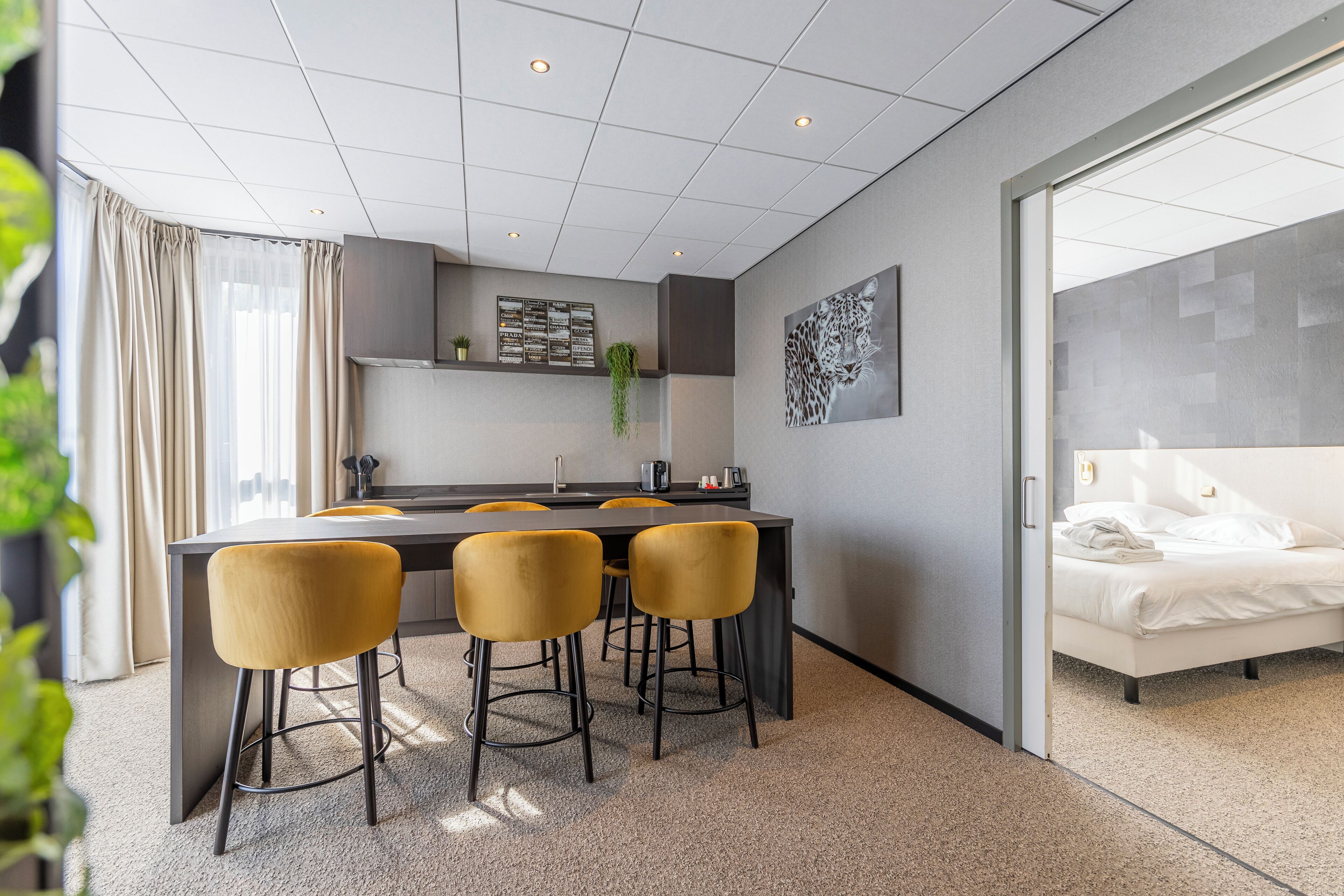 Une chambre d’hôtel moderne avec un grand lit, une petite kitchenette et un coin repas avec des chaises jaunes.