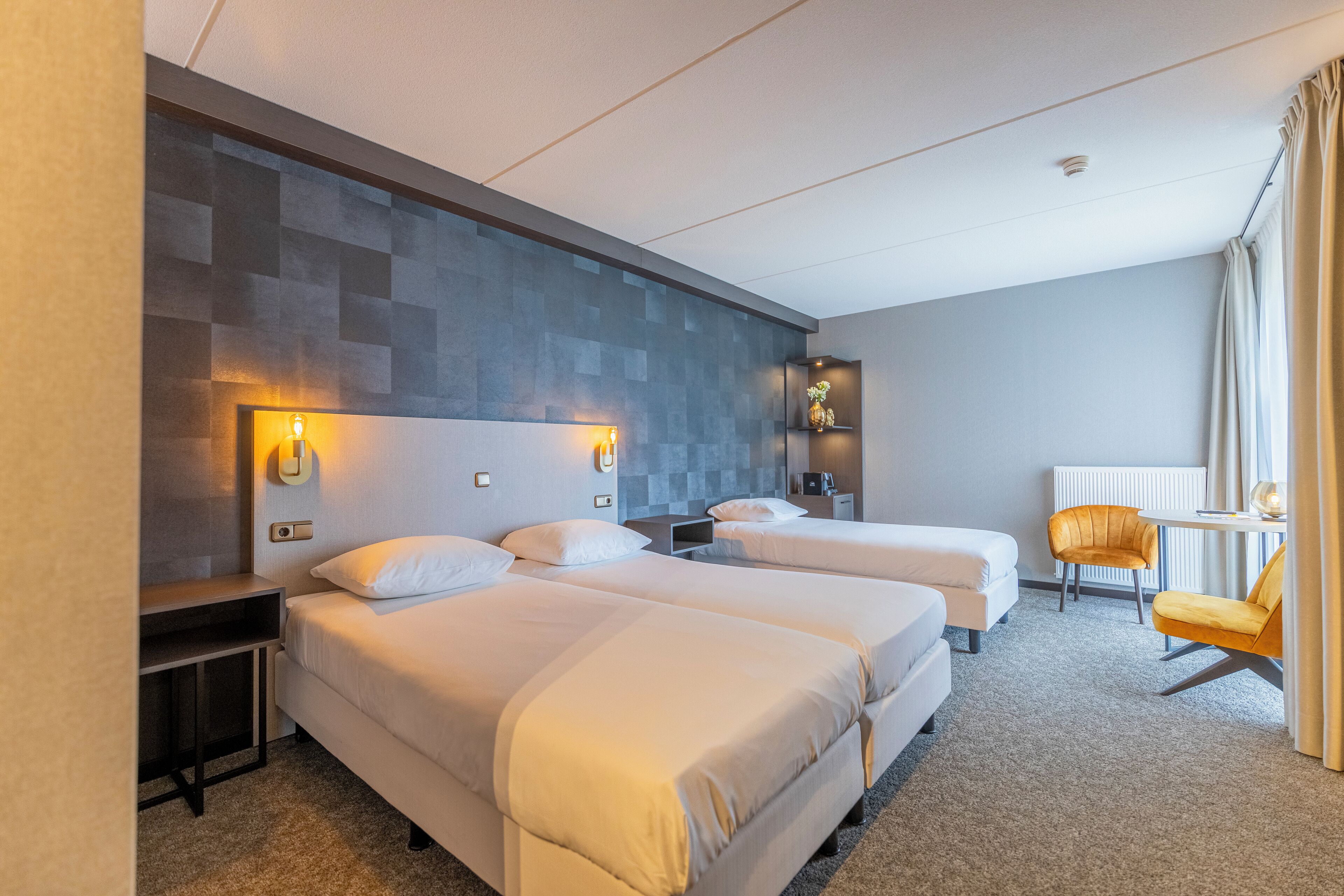 Een hotelkamer met twee bedden, een klein tafeltje, een stoel en een wandplank met een vaas.