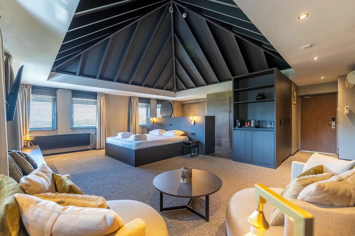 A modern hotel room with a large bed, a sitting area with a round table, and a kitchen area.