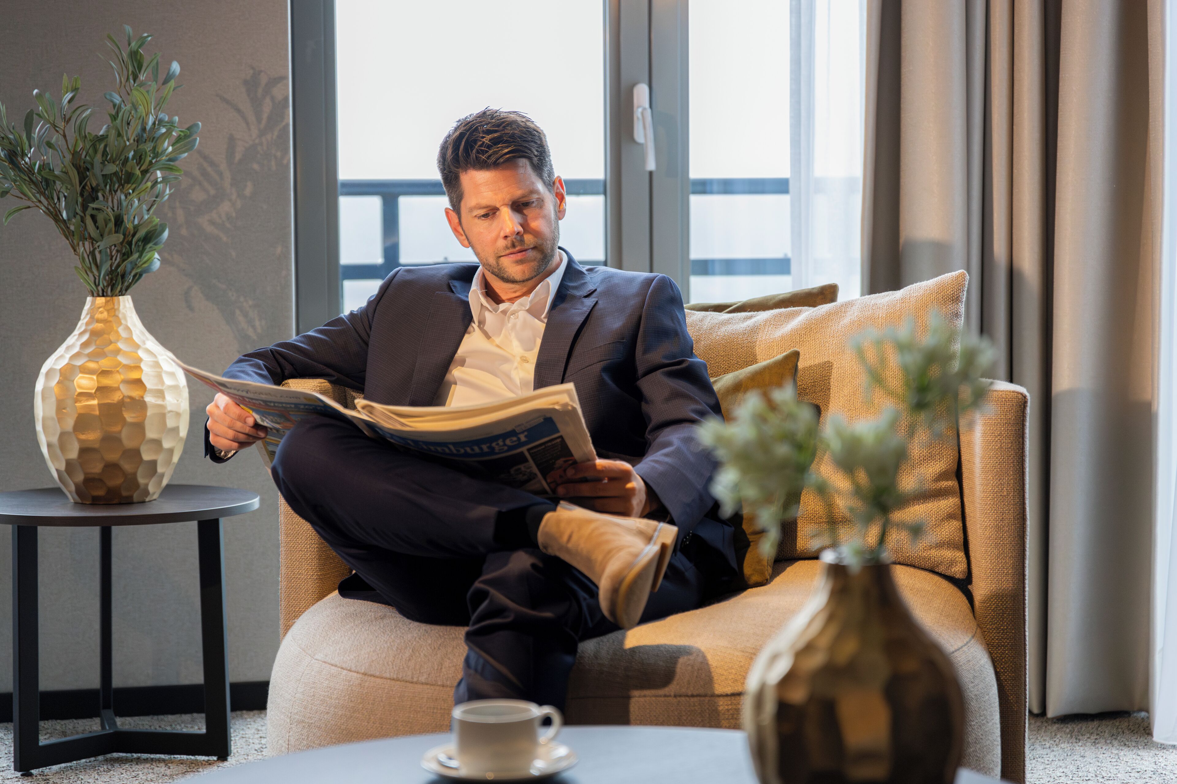 Een man zit op een bank een krant te lezen in een moderne kamer met een groot raam, een klein tafeltje met een kopje en decoratieve vazen.