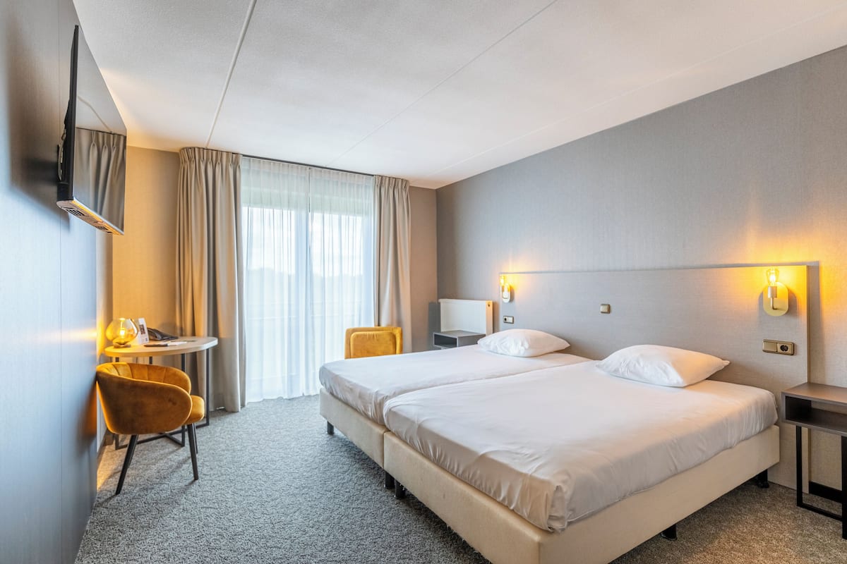 A hotel room with two beds, a desk with a chair, a TV mounted on the wall, and a window with curtains.