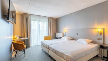 Ein Hotelzimmer mit zwei Betten, einem Schreibtisch mit Stuhl, einem an der Wand montierten Fernseher und einem Fenster mit Vorhängen.