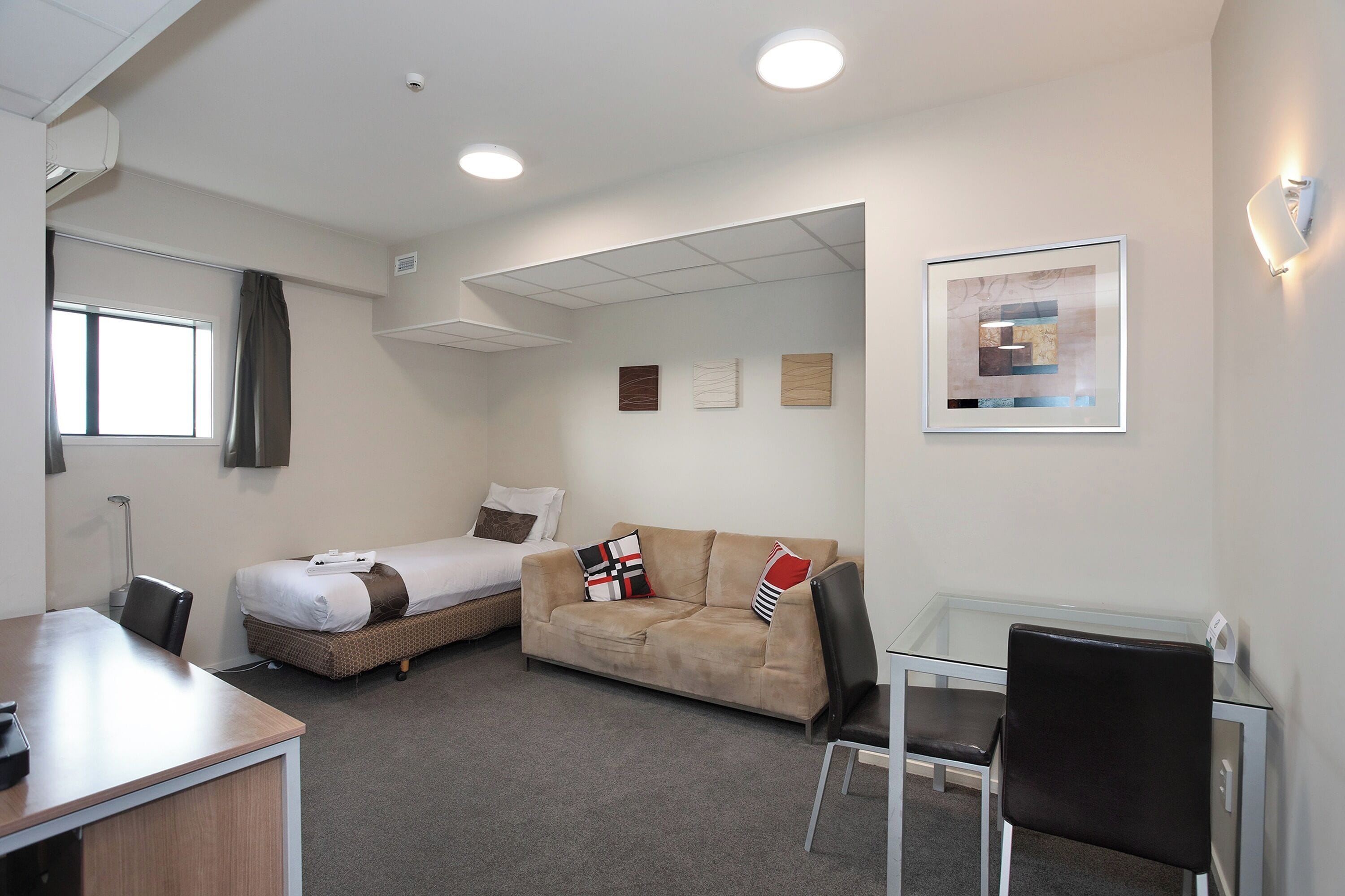 A hotel room with a bed, a sofa, a desk, and a chair. There is a window with curtains, a ceiling light, and wall art.