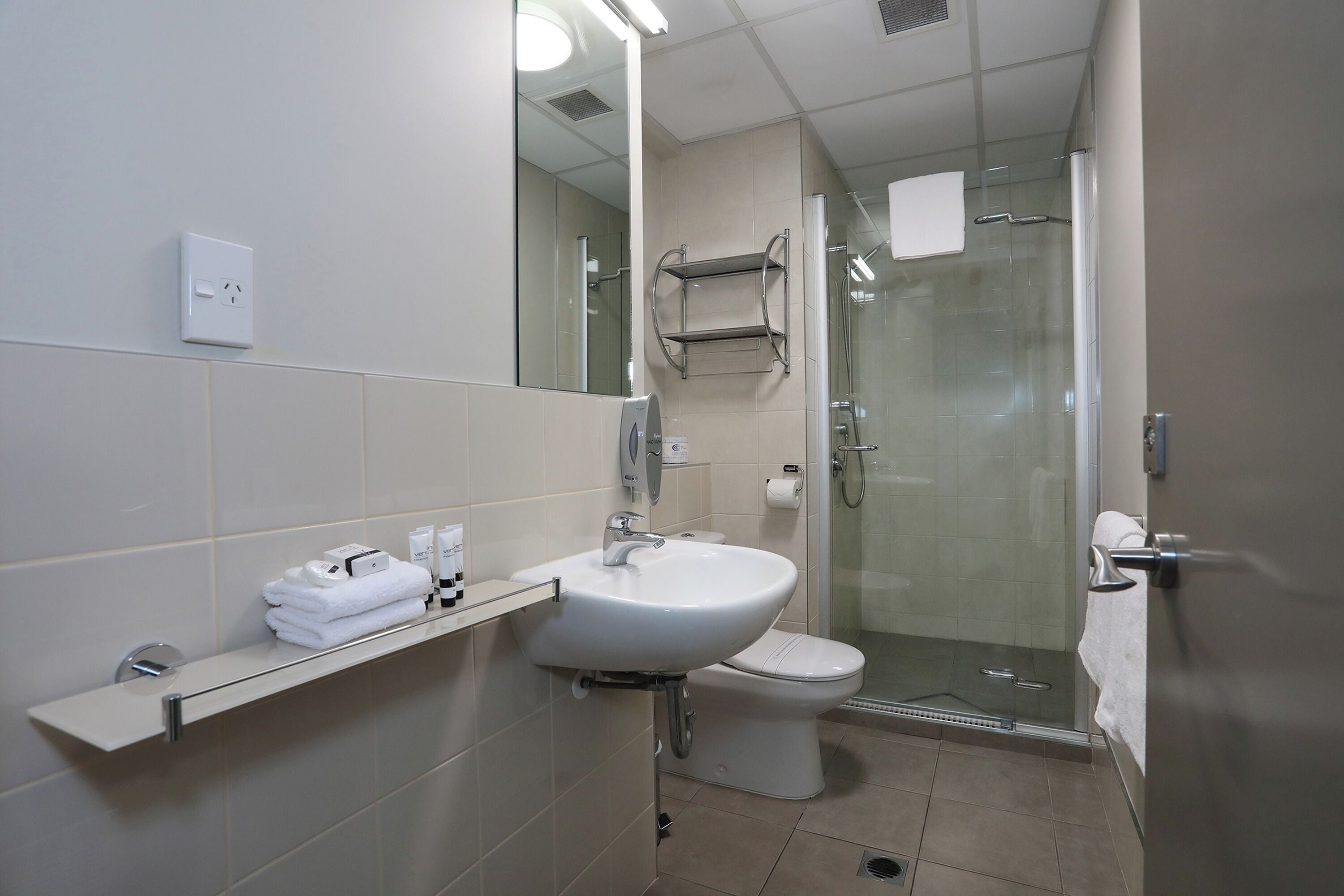A modern bathroom with a white sink, toilet, and shower. There is a towel rack and a shelf with toiletries.