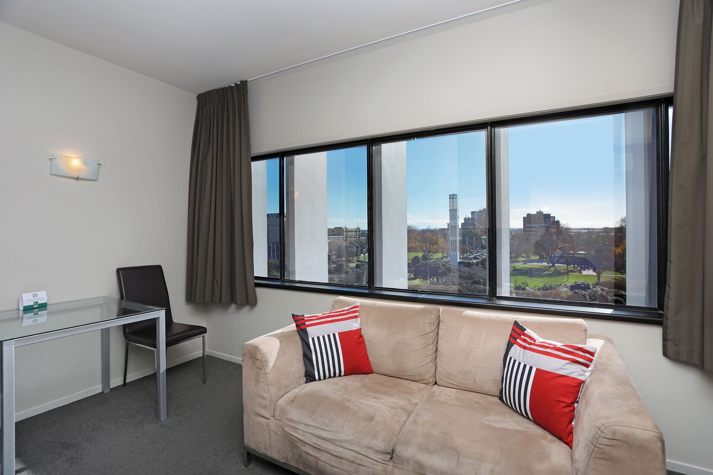 Una habitación moderna con un escritorio de vidrio, un sofá color crema con almohadas rojas y blancas, y un ventanal con vistas a la ciudad.