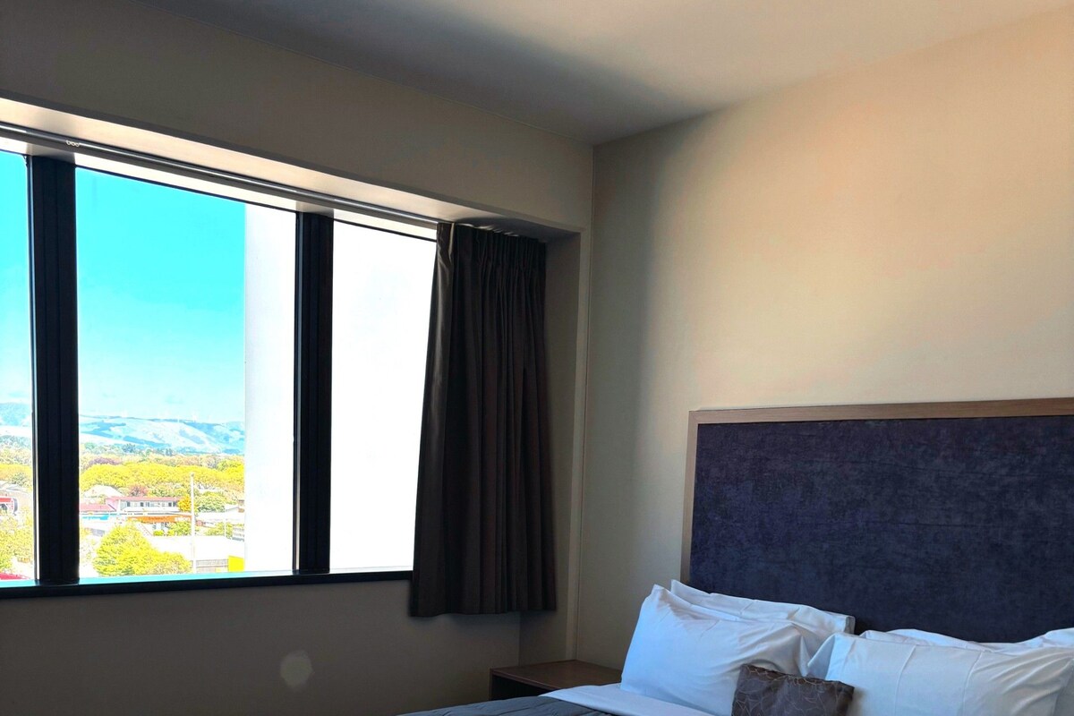 A hotel room with a bed, a nightstand, a window with curtains, and a view of a landscape.