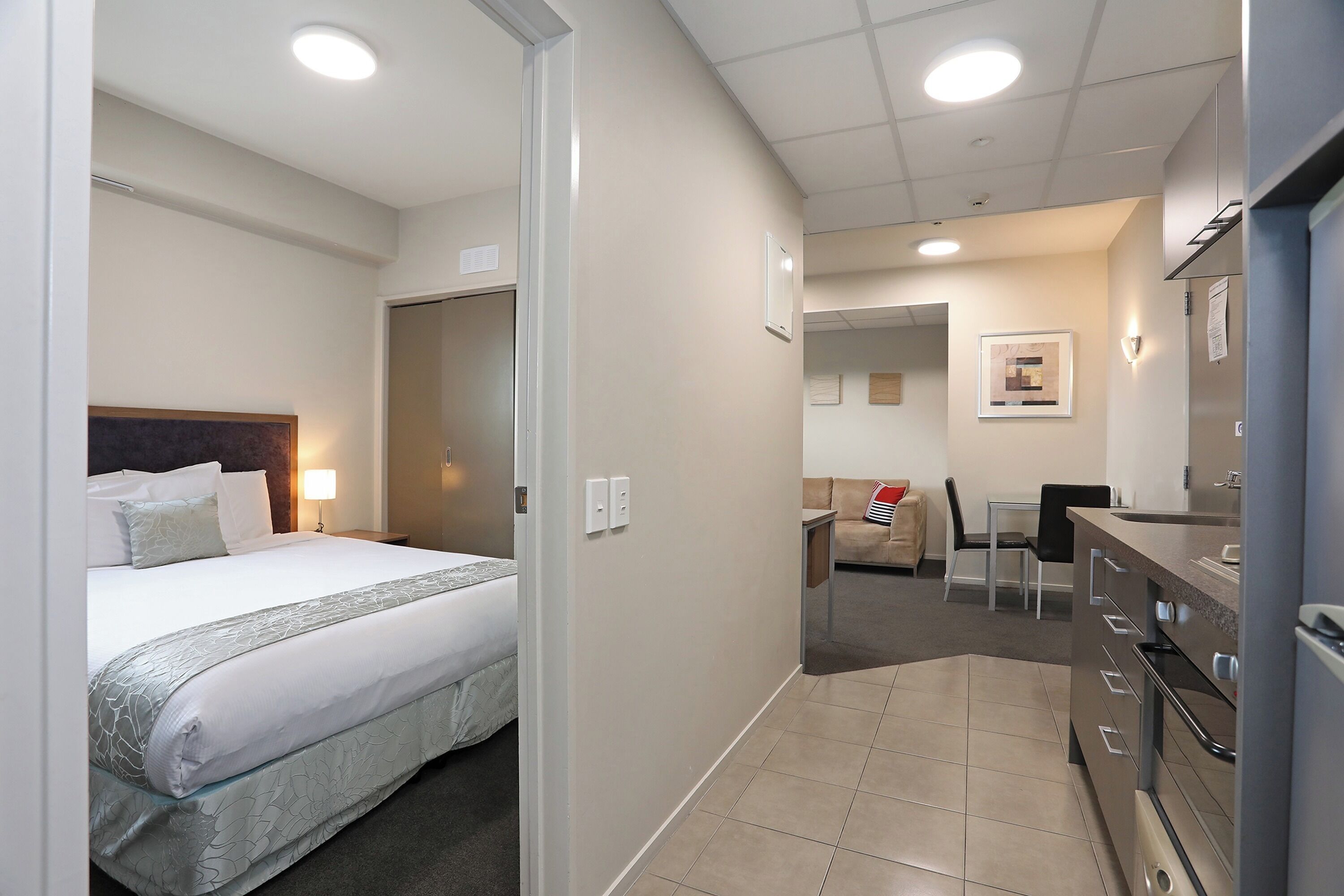 A hotel room with a bed, bedside table, lamp, and a view of the hallway leading to the bathroom and kitchen area.