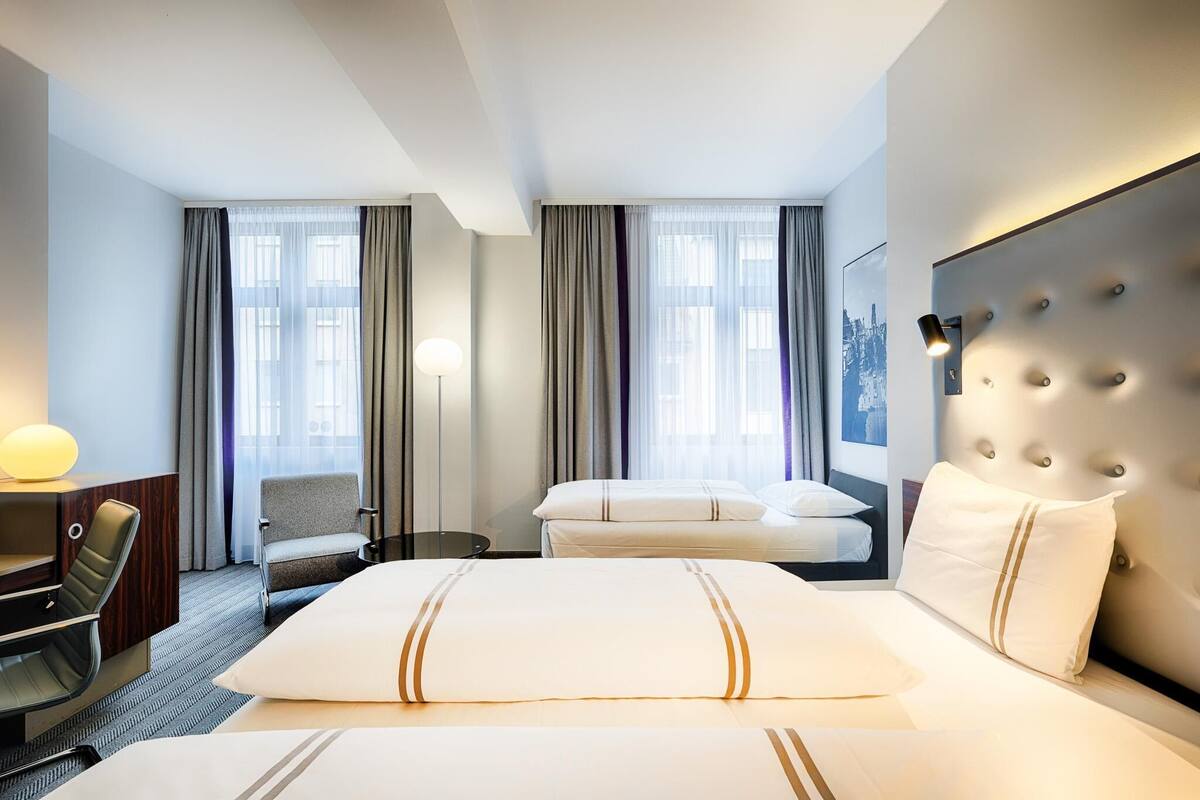 A hotel room with a large bed, a desk with a chair, a sofa, and a headboard with a light fixture.