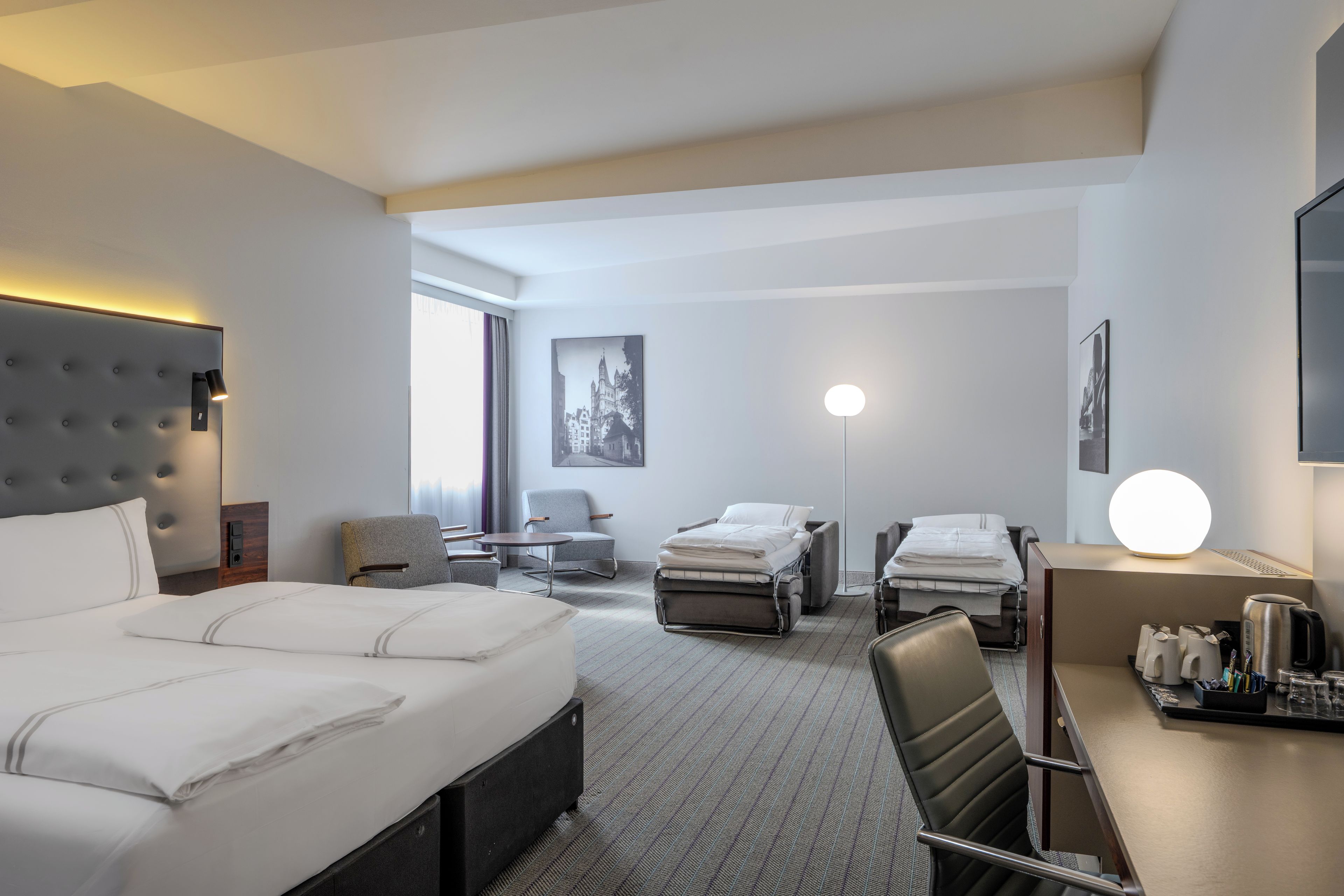 A hotel room with a large bed, two smaller beds, a desk, a chair, a flat-screen TV, and a large window.