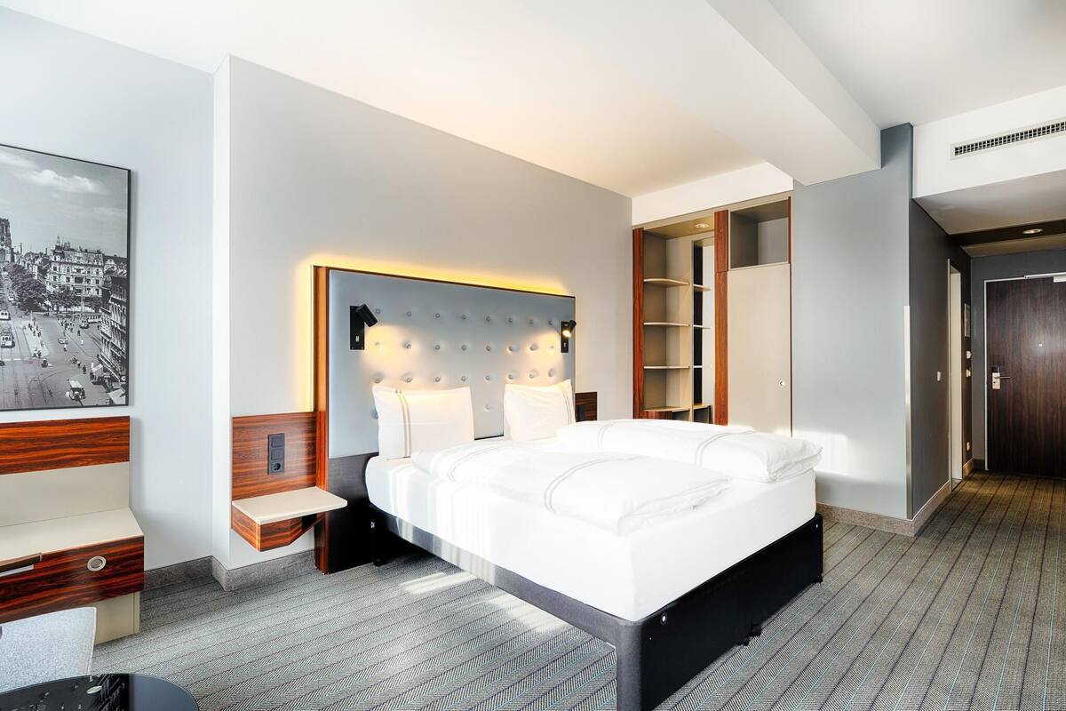 A modern hotel room with a large bed, bedside tables, a built-in shelf, and a framed cityscape picture.