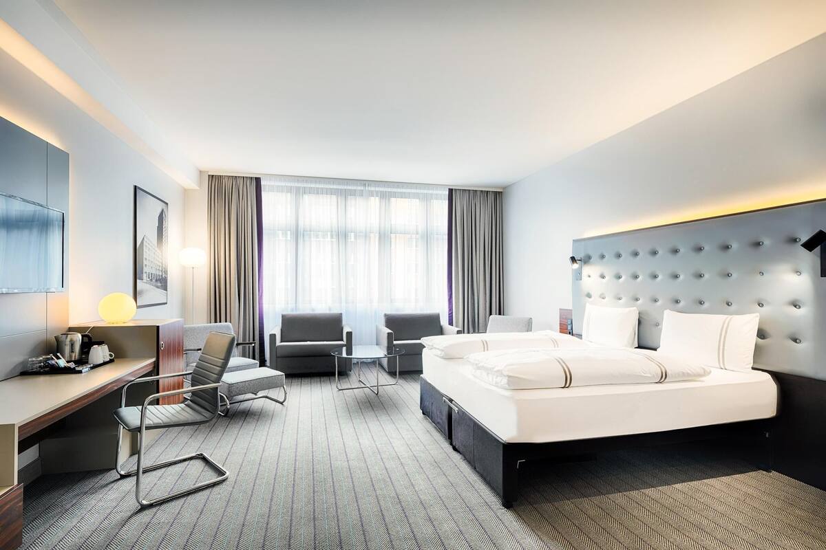 A hotel room with a bed, two armchairs, a desk, a lamp, and a large window with curtains.