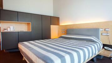 A modern hotel room with a large bed, a built-in wardrobe, a bedside table, and a small shelf with toiletries.