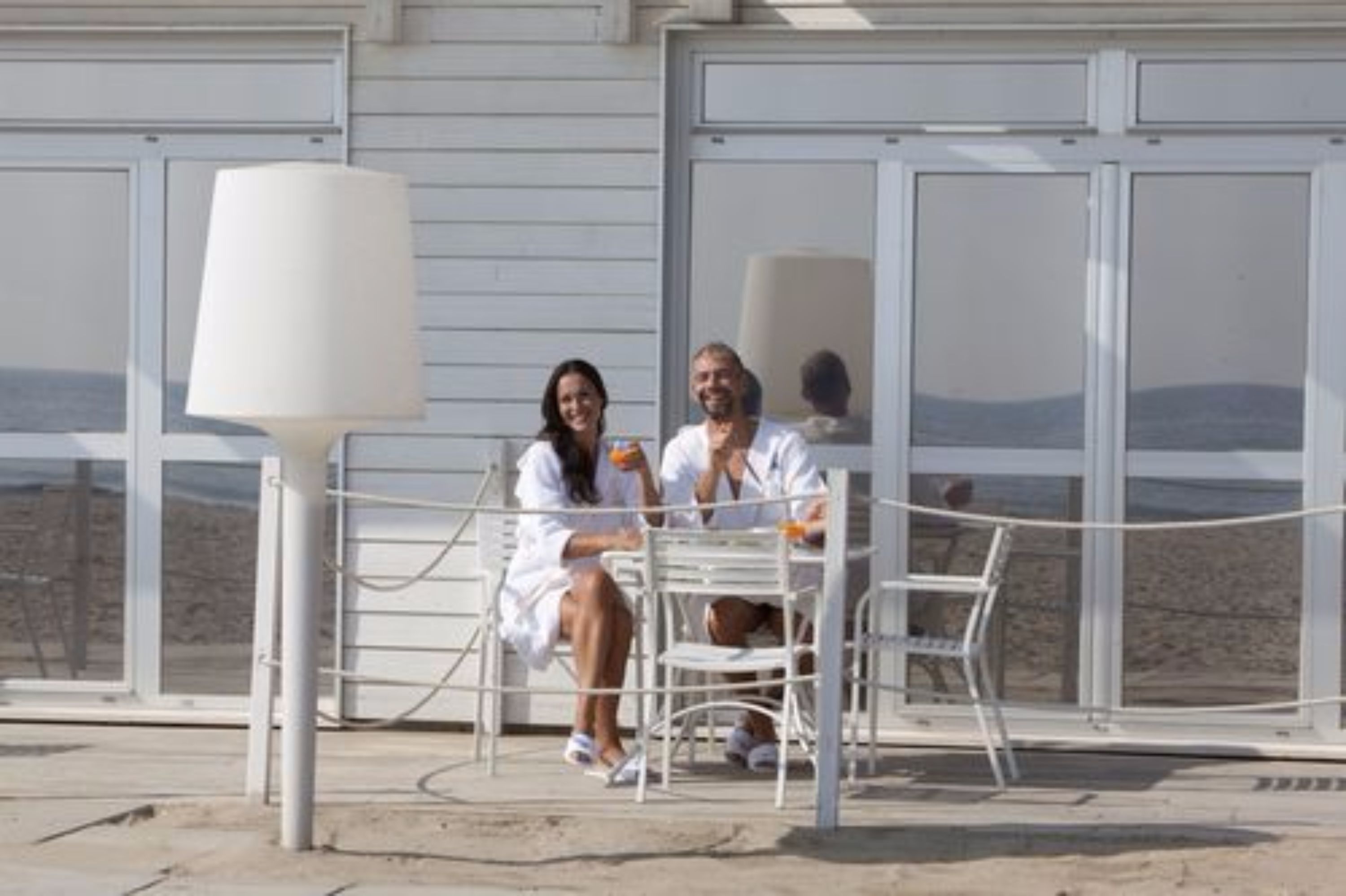 Ein Paar sitzt an einem Tisch mit Blick auf den Strand, eine weiße Lampe und ein Geländer sind zu sehen.