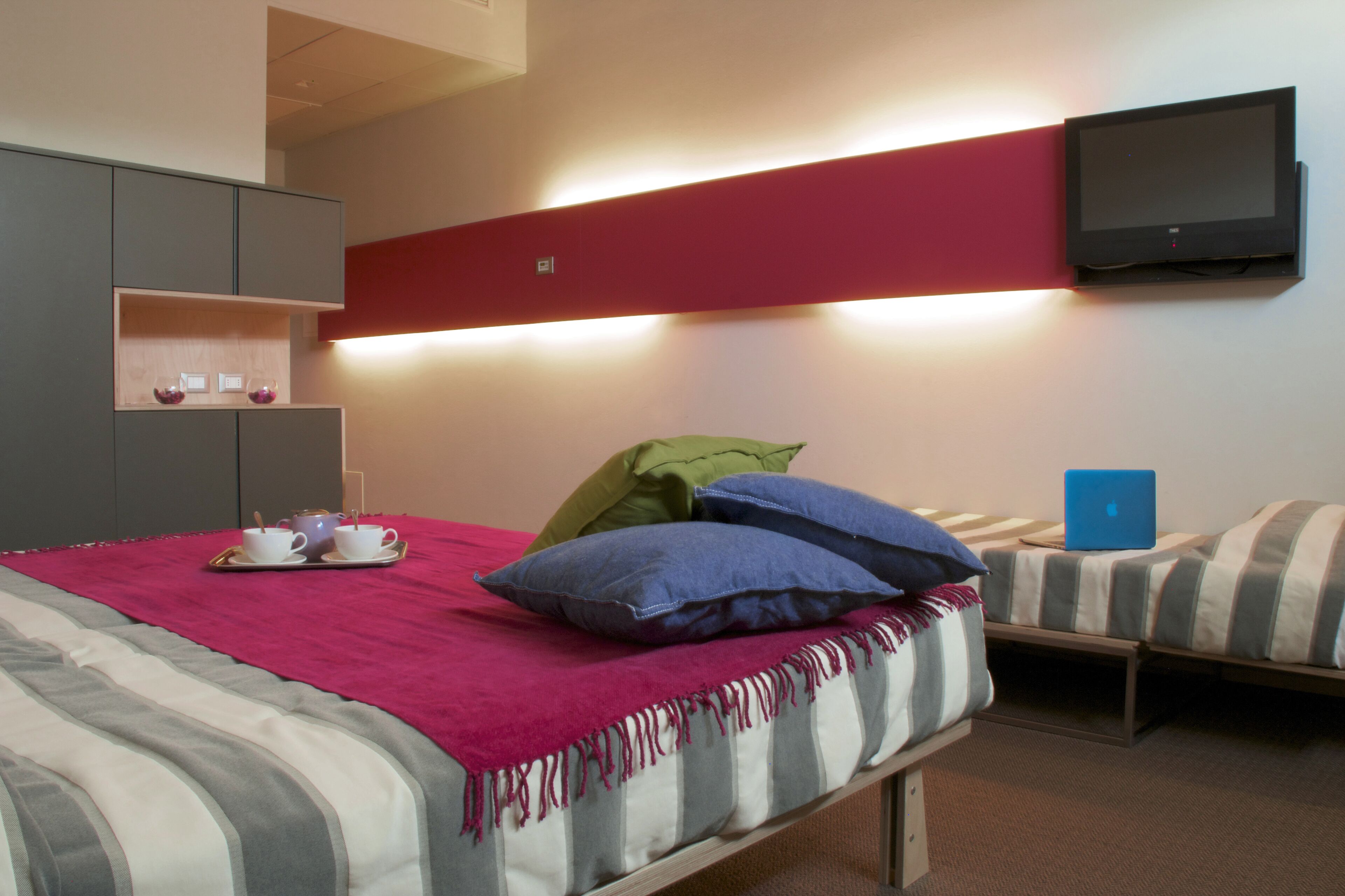 A hotel room with a bed, a TV mounted on the wall, a laptop on a bedside table, and a shelf with cups and a spoon.