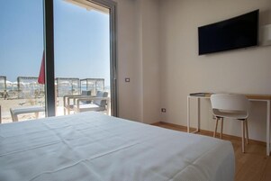 A hotel room with a bed, a desk, a chair, a TV, and a balcony with a view of the beach.