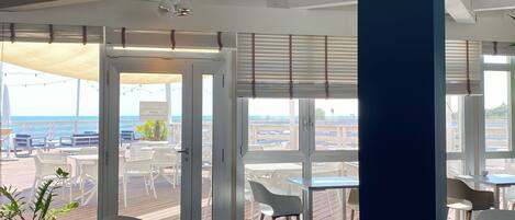 A modern interior with a view of the sea, featuring a dining area with tables and chairs, large windows, and a blue accent wall.