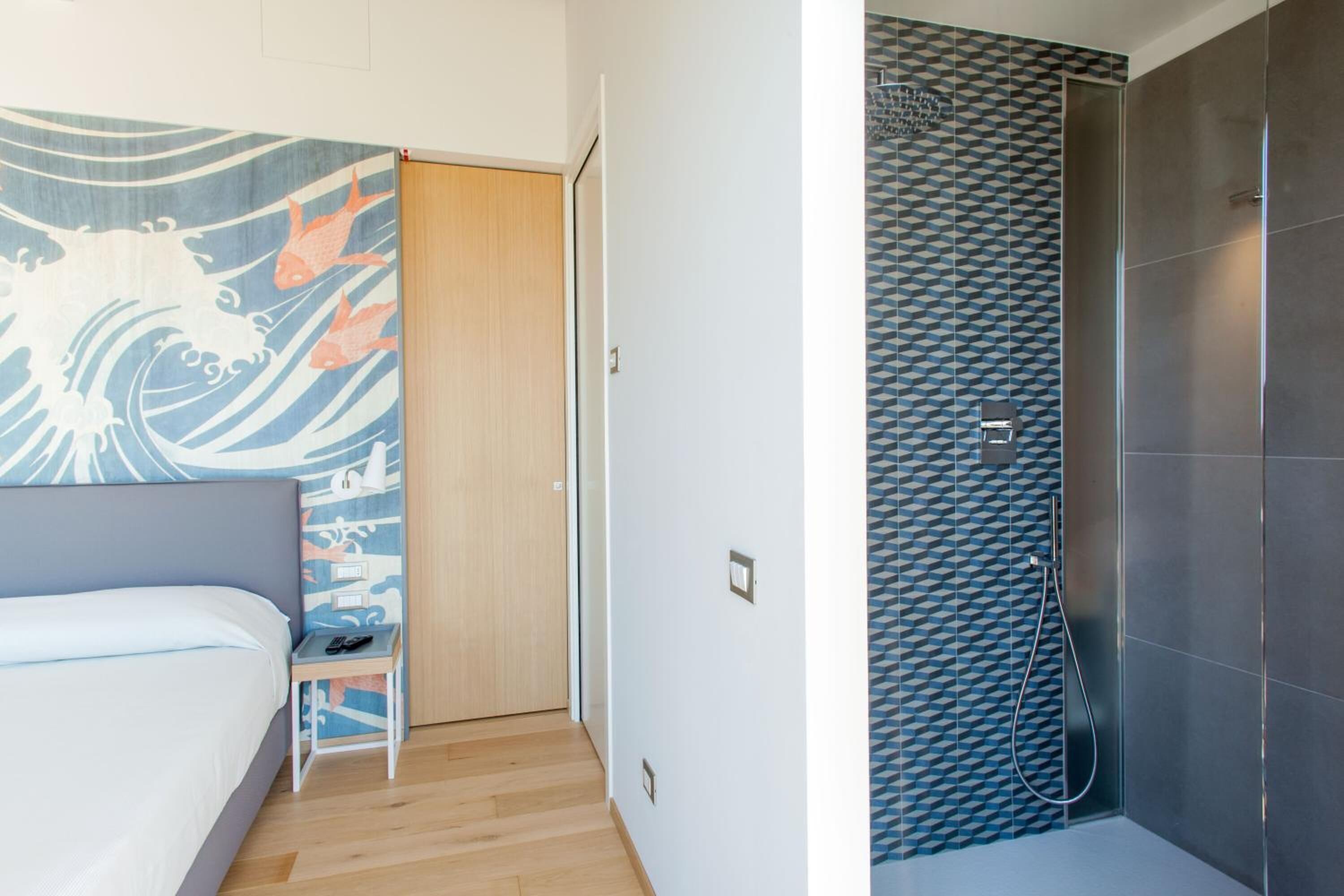 A bedroom with a bed, a nightstand, and a wall with a wave pattern. The room has a wooden door and a tiled shower area with a geometric pattern.