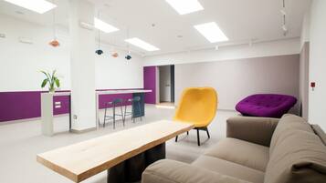 Ein moderner Arbeitsraum mit einem langen Tisch, einem gelben Stuhl, einem violetten Stuhl und einem beigen Sofa.