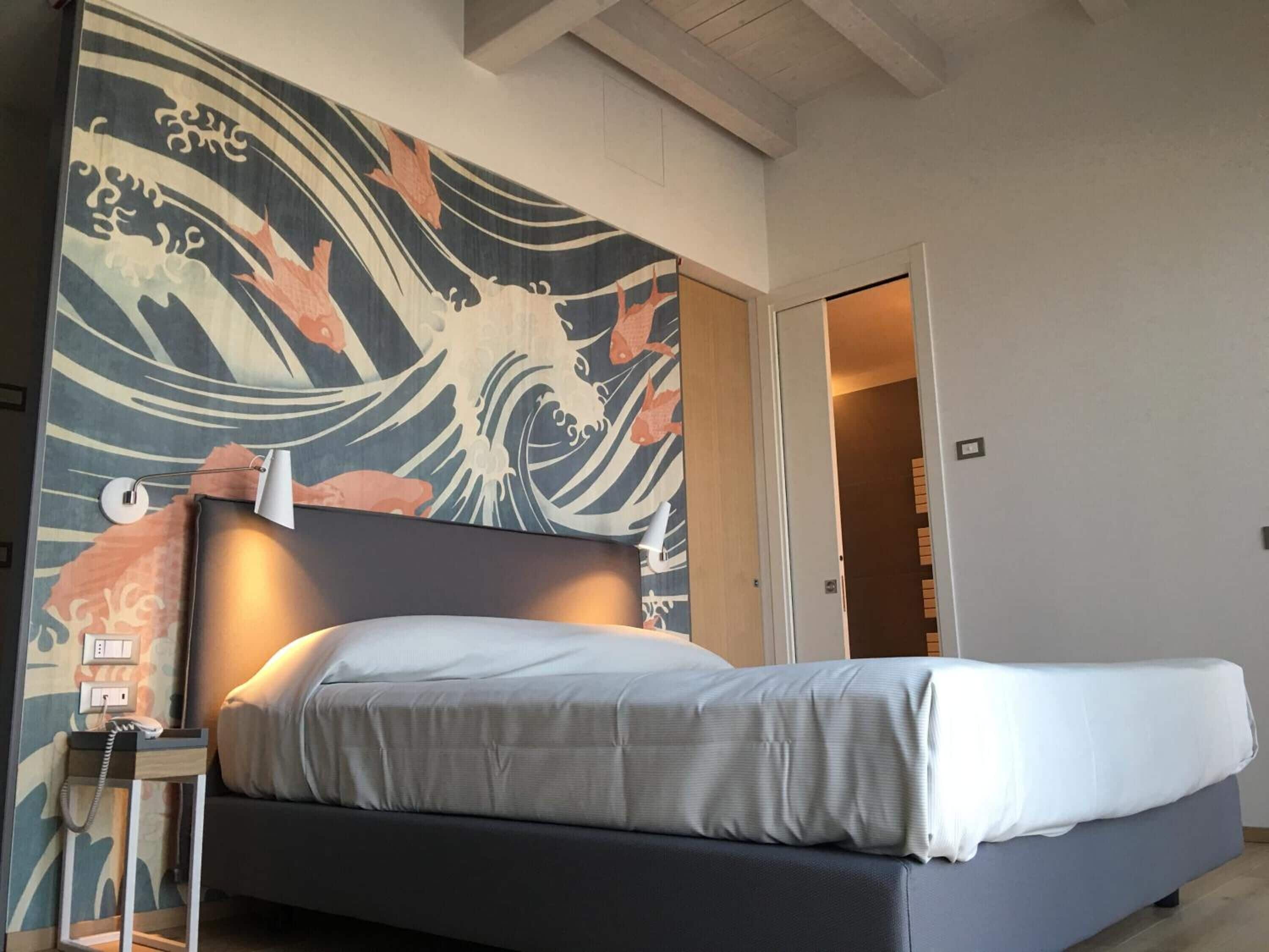 A modern hotel room with a large Japanese wave painting on the wall, a bed with a grey headboard, and a bedside table with a lamp.