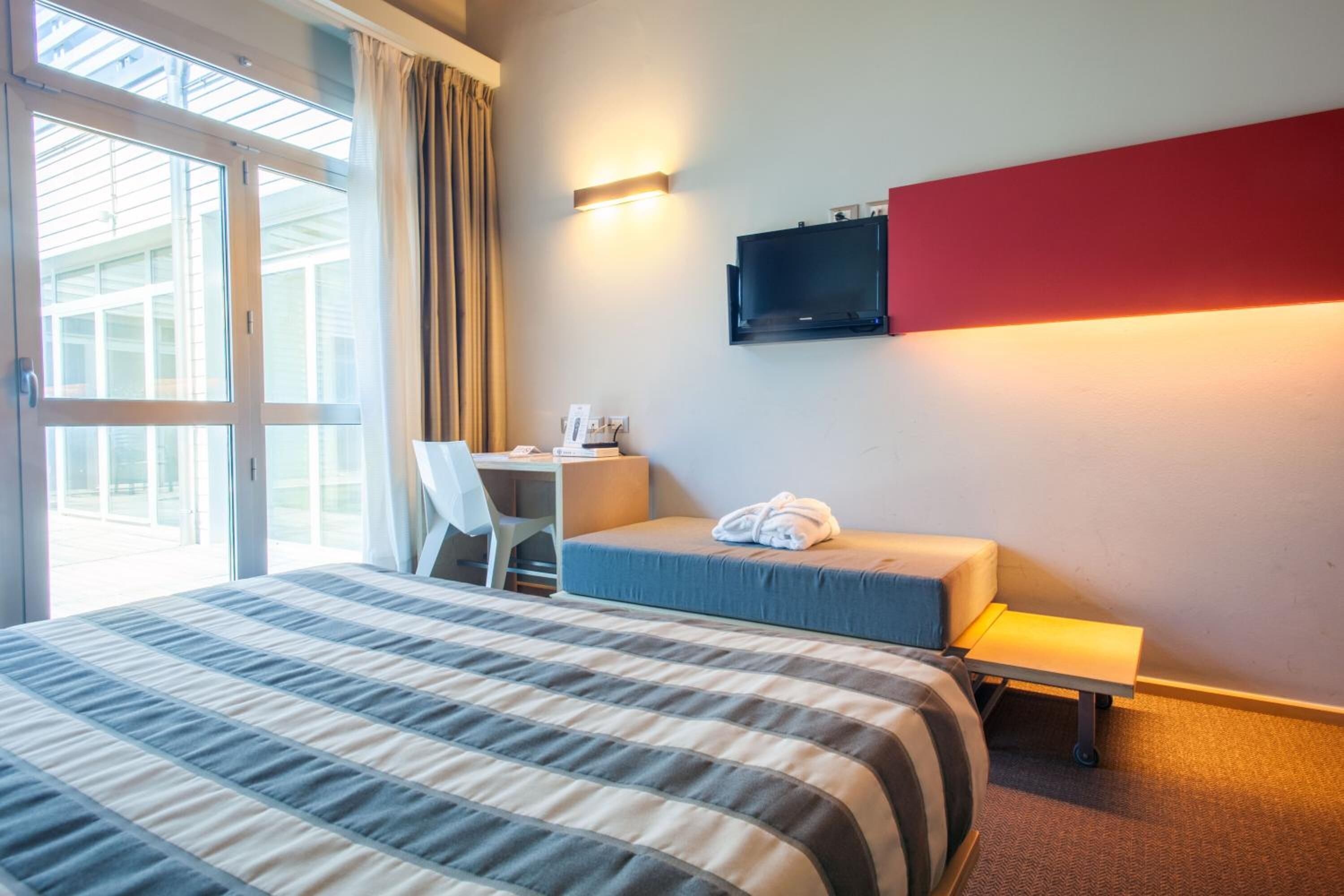 Une chambre d’hôtel avec un grand lit, un bureau avec une chaise, un téléviseur fixé au mur et une vue sur l’extérieur, accessible par une porte vitrée.