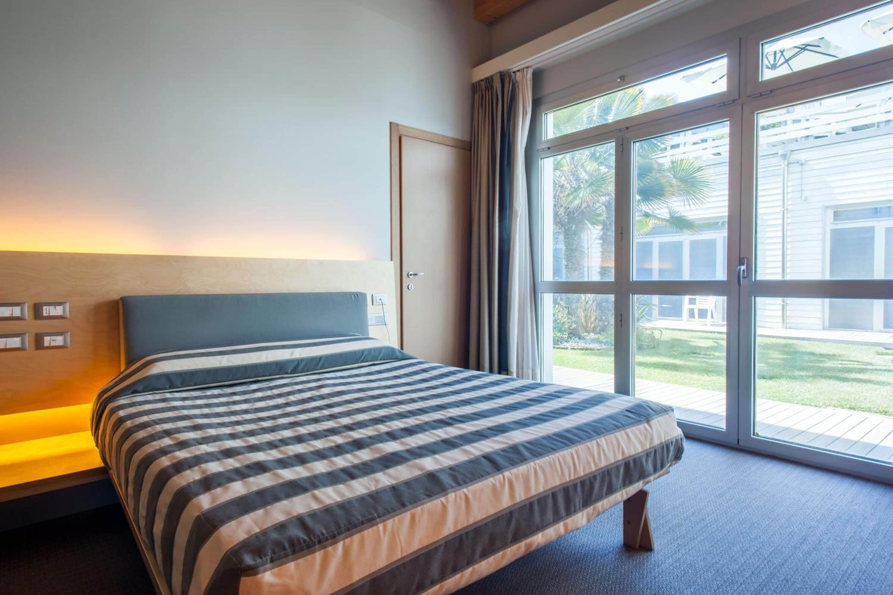 A hotel room with a bed, a bedside table, a window with curtains, and a view of the outdoors.