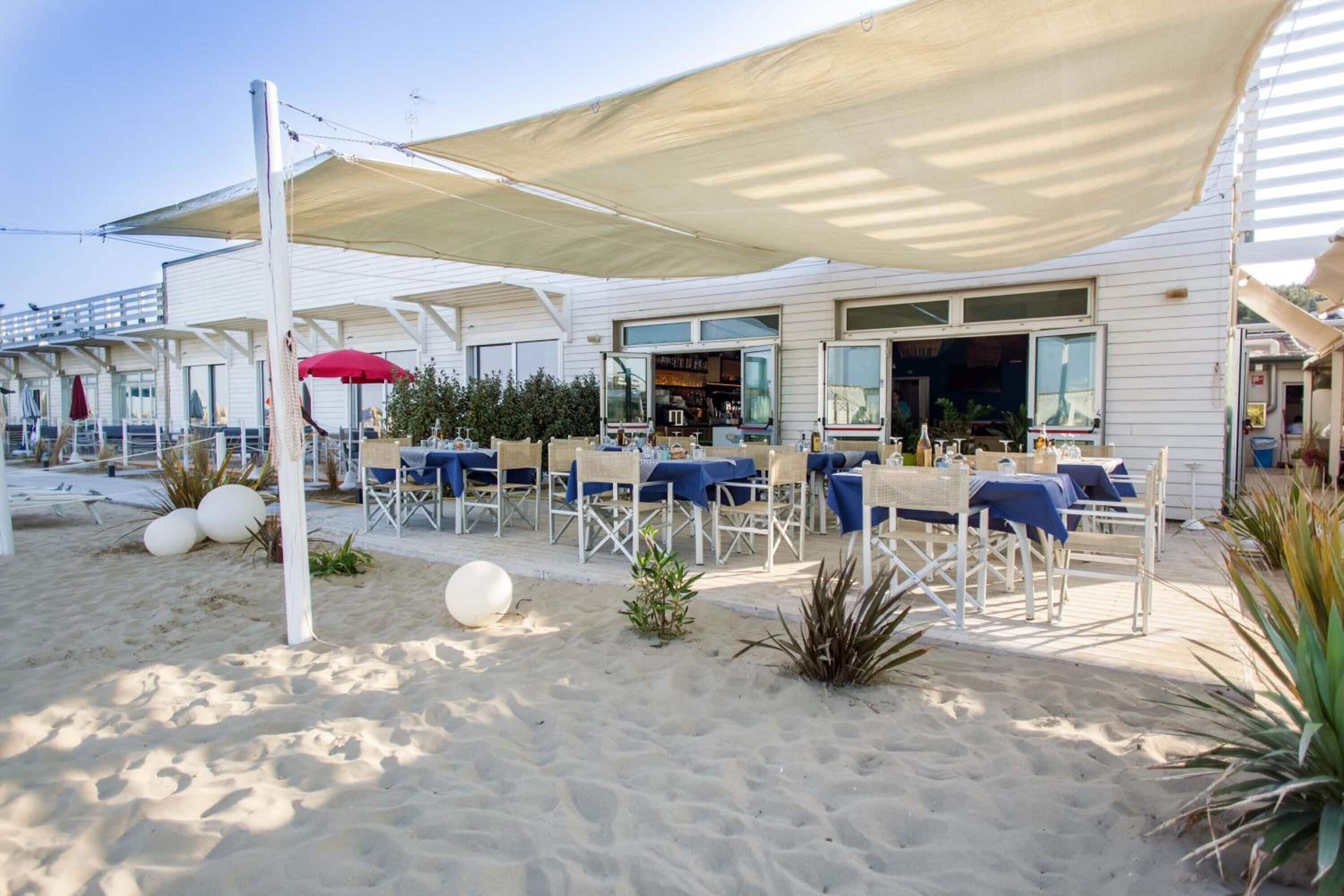 An outdoor seating area with tables and chairs under a canopy, a beach setting with sand and plants, and a building with a bar visible through the windows.