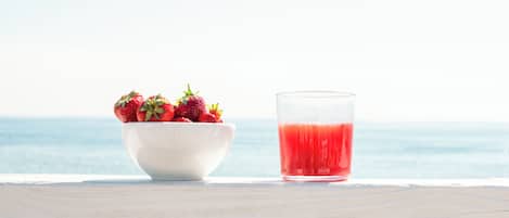 A white bowl of strawberries and a glass of red liquid on a table by the sea.