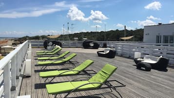 Una terraza con sillones reclinables verdes y un sofá negro, con vistas a una playa y un área boscosa.