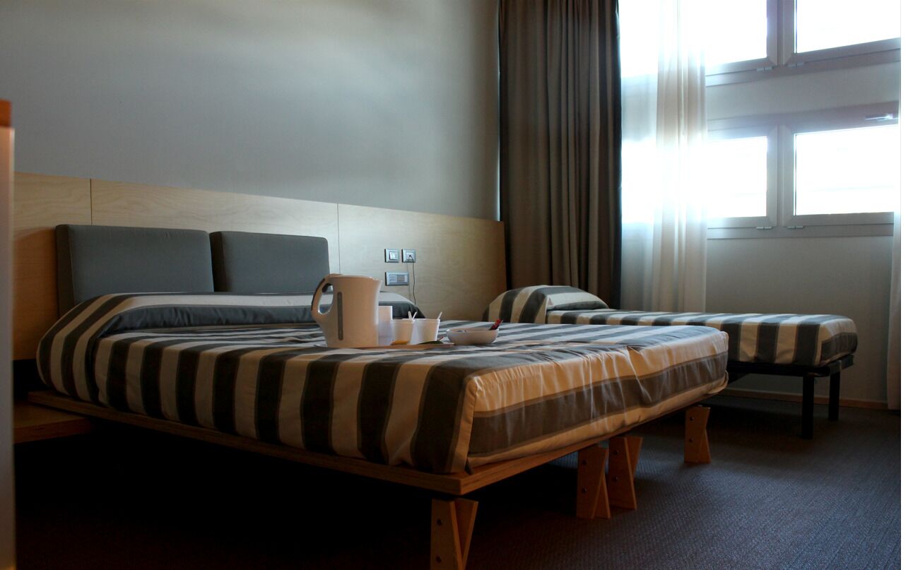 Camera d'albergo con due letti, una testiera in legno, una finestra con tende e un comodino con lampada e tazza.