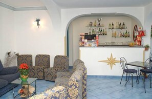 A bar area with shelves of bottles and snacks, a star decoration on the wall, and a seating area with patterned sofas and a table with chairs. - Il Gabbiano (Positano)