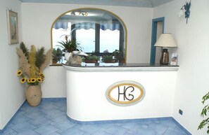 A reception area with a curved counter, a mirror above it, and a potted plant with sunflowers. - Il Gabbiano (Positano)