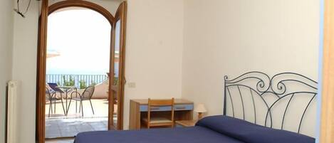 A bedroom with a bed, a desk, and a chair, an open door leading to a balcony with a view.