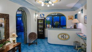 A reception area with a blue-tiled floor, a reception desk with a logo, a chandelier, and a view of the outside through a window. - Il Gabbiano (Positano)
