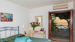 A bedroom with a bed, a desk with a vase of flowers, a painting on the wall, and a view of the sea through an open door. - Il Gabbiano (Positano)